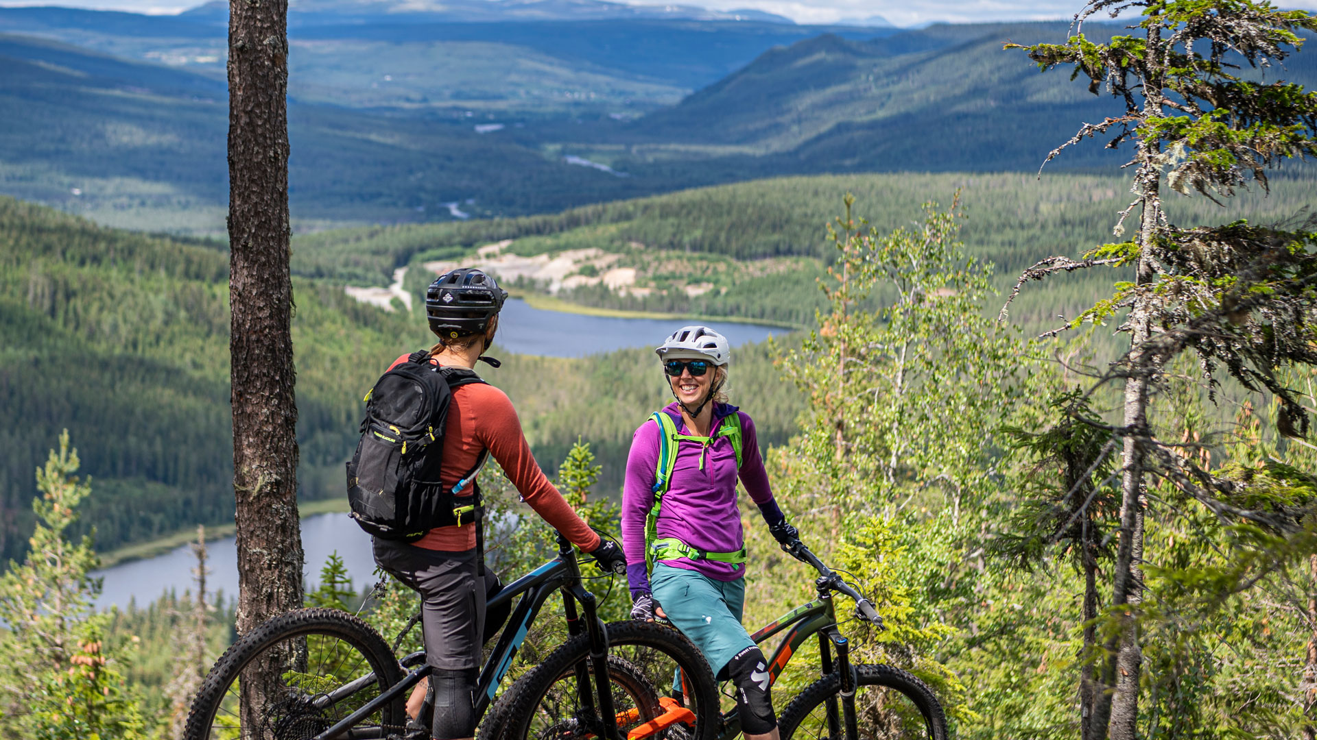 Två mountainbikecyklister på en skogsstig med utsikt över en sjö och berg i bakgrunden, sommarlandskap i Sverige