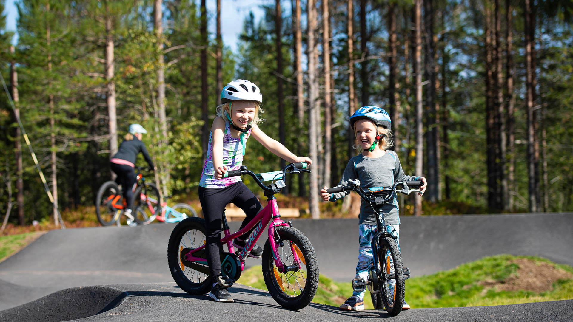 Barn cyklar i en pumptrack i skogen, med cykelhjälmar på, omgivna av tallar och natur