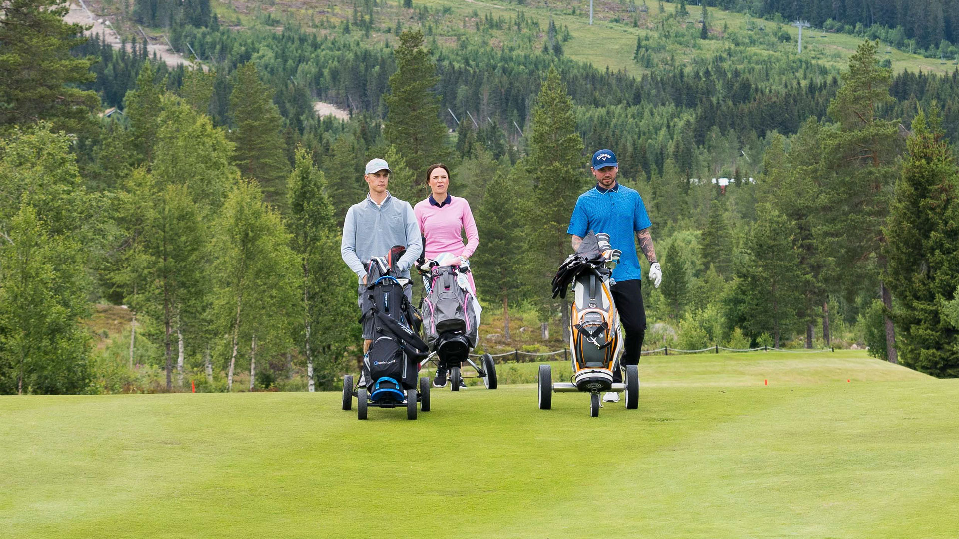 Tre personer spelar golf på en grön golfbana omgivna av skog och berg