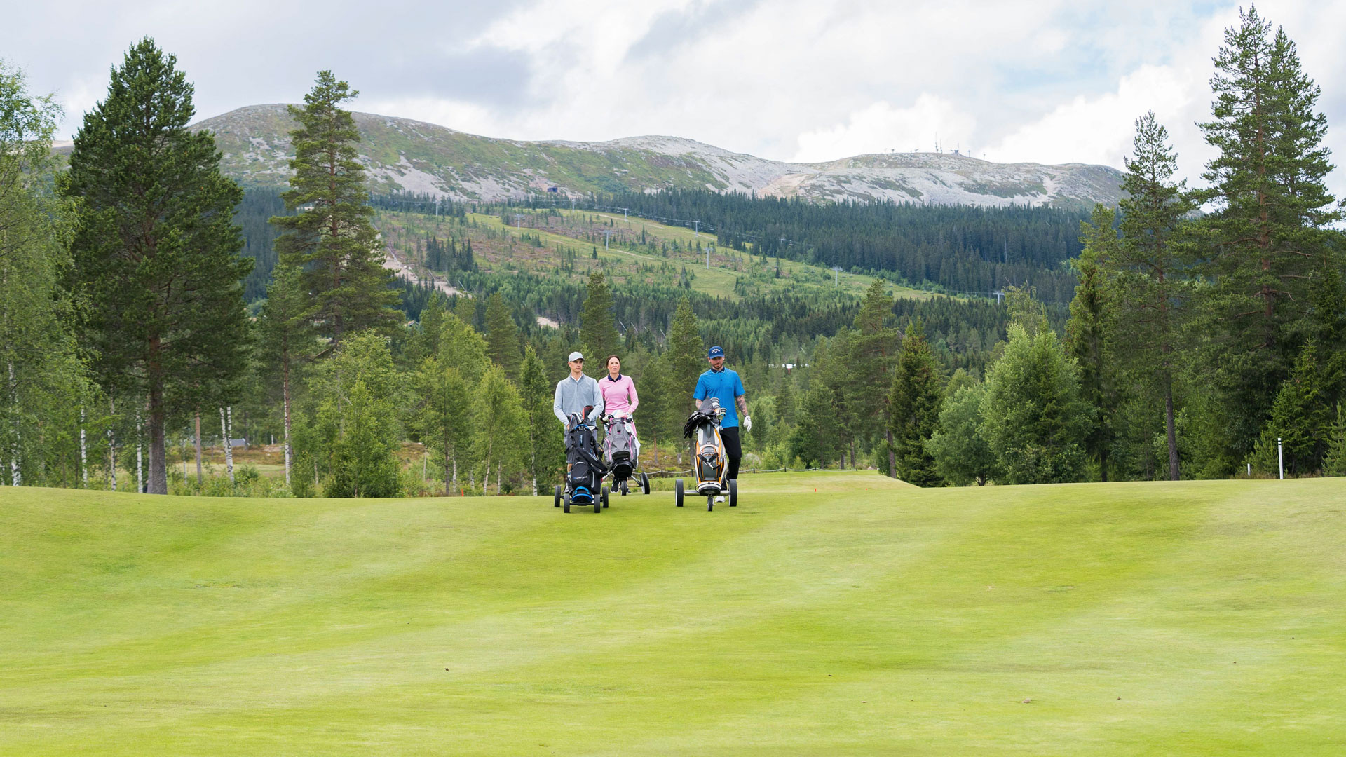Tre personer spelar golf på en grön golfbana omgiven av skog och berg i fjärran, under en molnig himmel