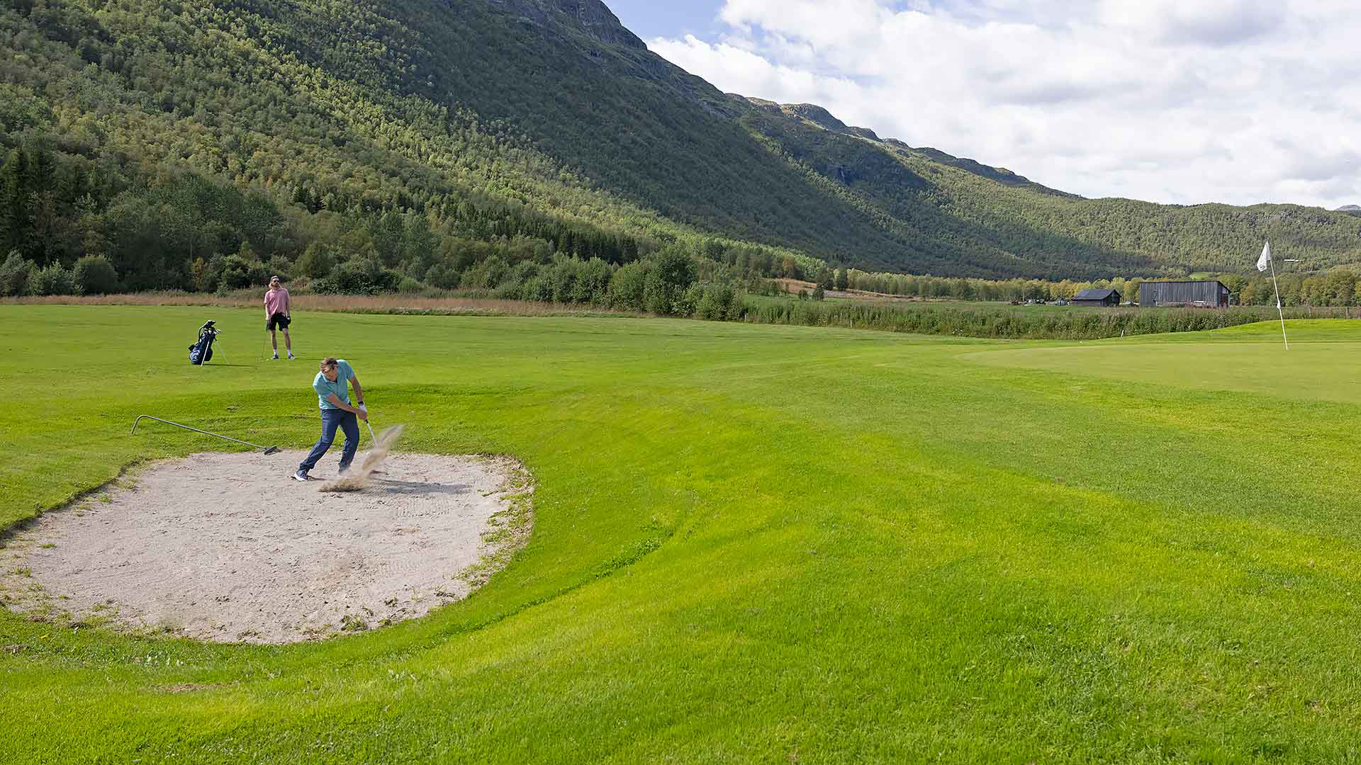 Golfspelare slår ett bunkerslag på en grön golfbana omgiven av berg och skog under en klar himmel