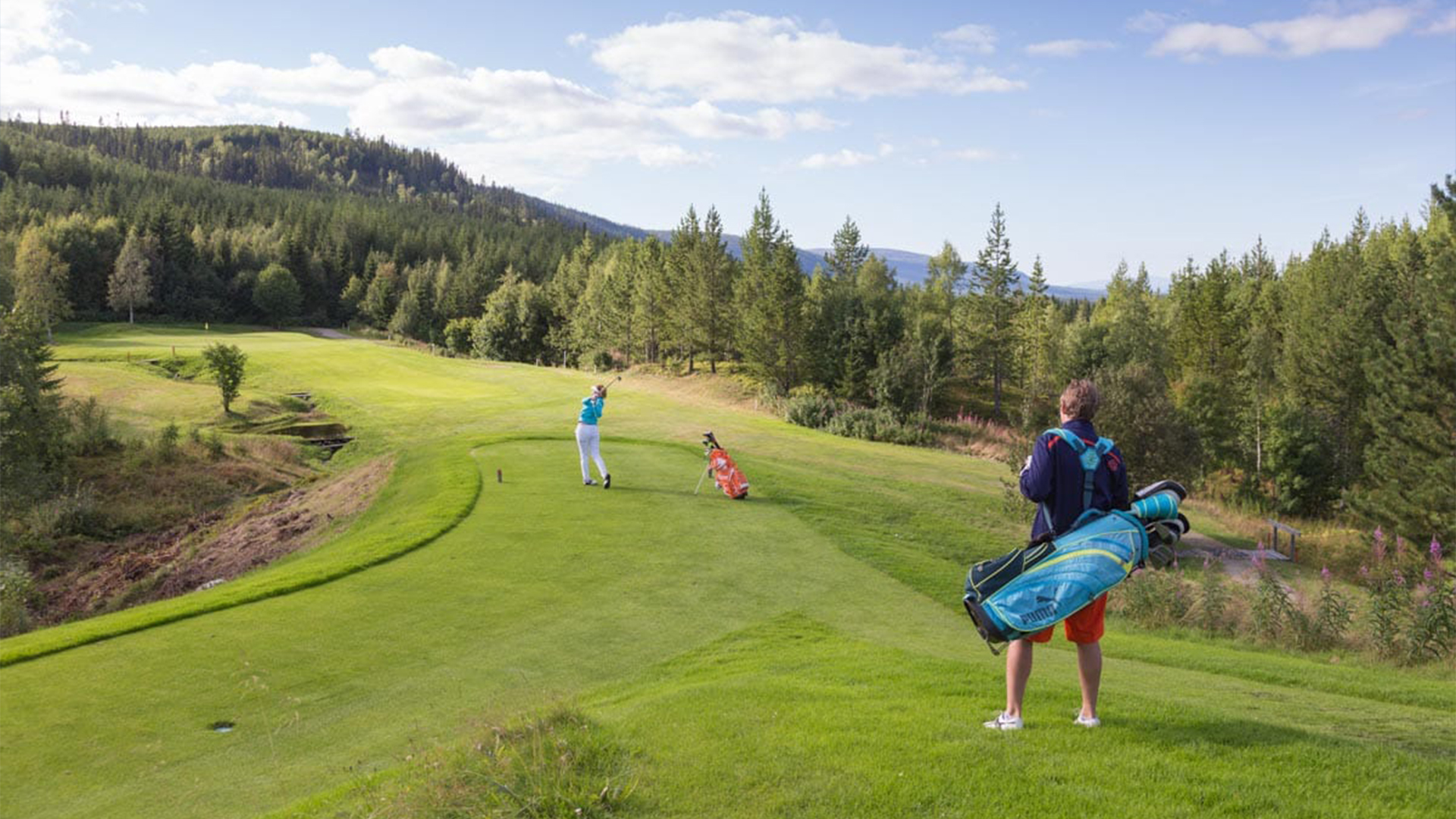 Personer som spelar golf på en grön golfbana omgiven av skog och berg i bakgrunden. Klarblå himmel med moln