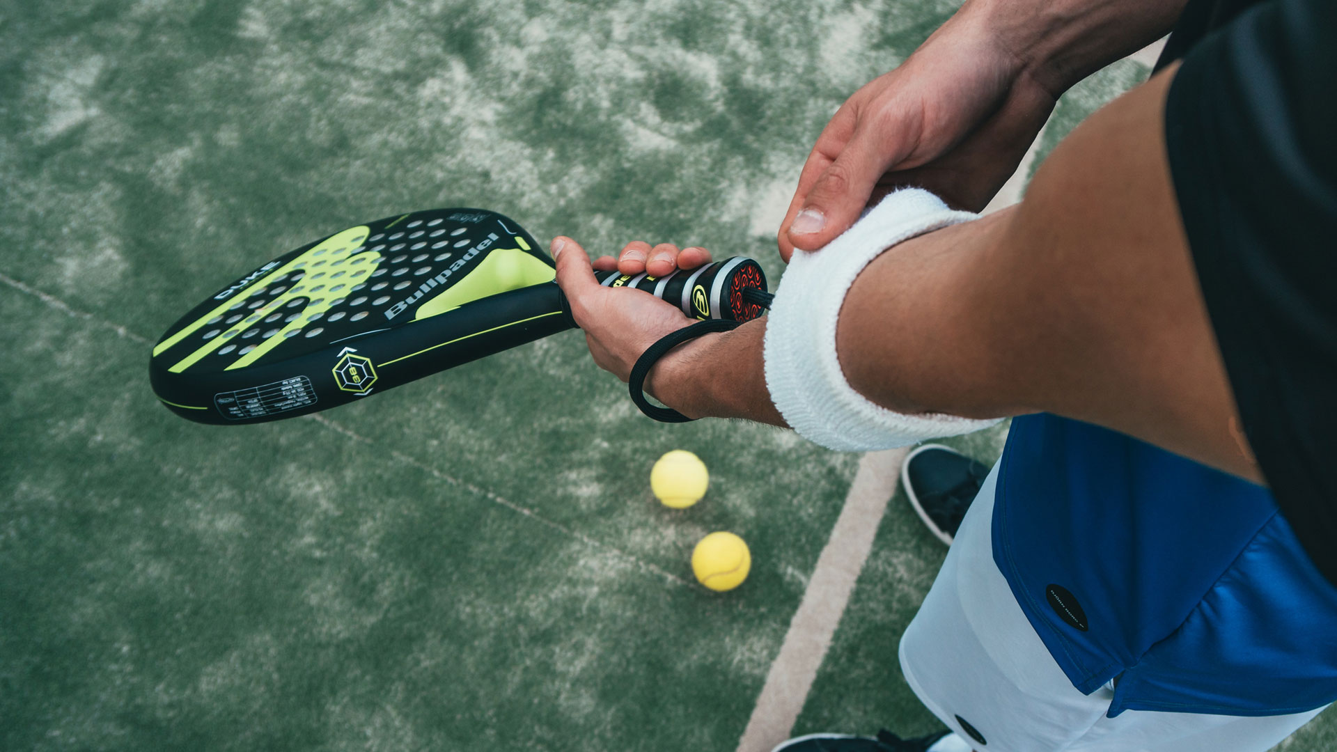 Person som förbereder sig för att spela padel, justerar sitt handledsband bredvid en padelracket och två bollar på en konstgräsplan