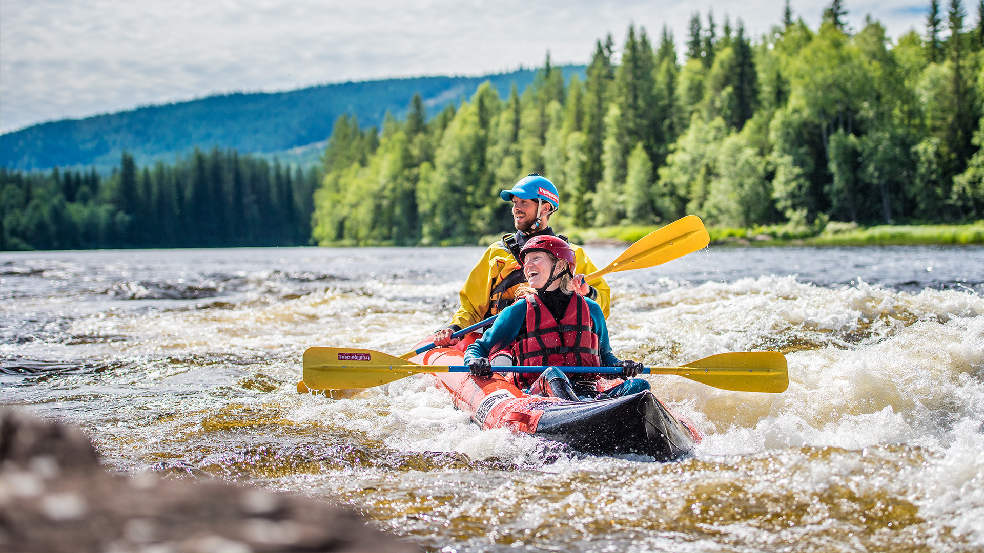 Två personer paddlar en kajak i forsande vatten omgivna av grön skog och berg i bakgrunden, njuter av en spännande friluftsupplevelse i Sverige