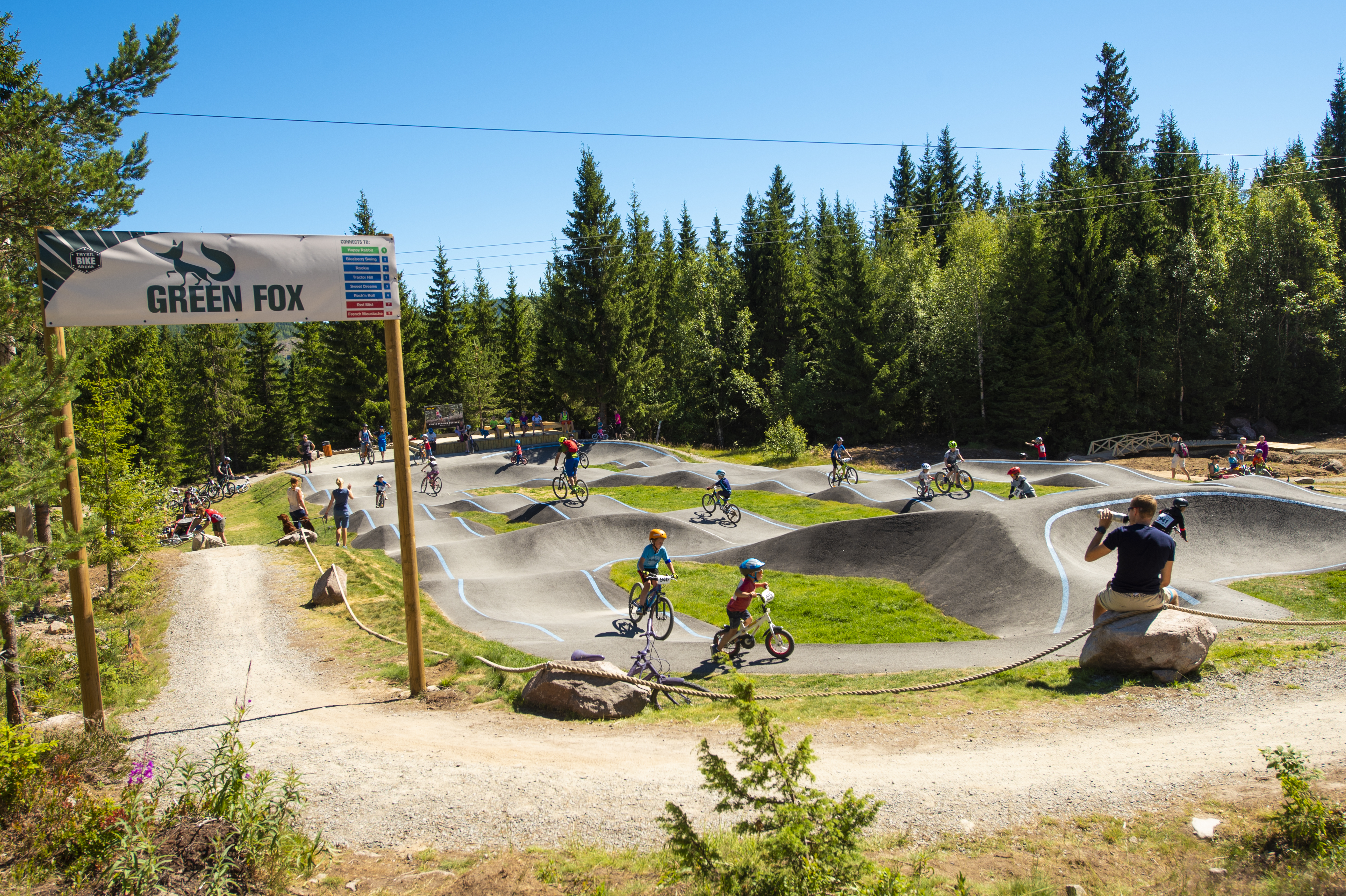 Cykelpark med pumptrack och cyklister omgivna av skog. Solig dag med klarblå himmel