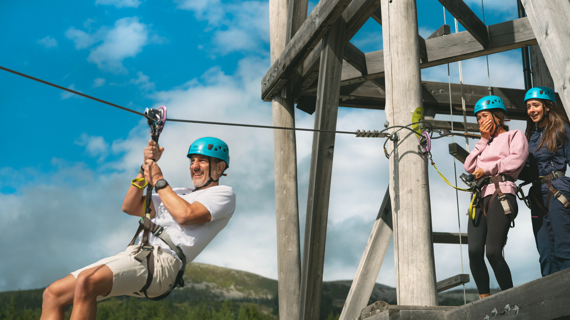 Man och två kvinnor med hjälmar upplever zipline-äventyr från en träplattform, omgivna av blå himmel och berg