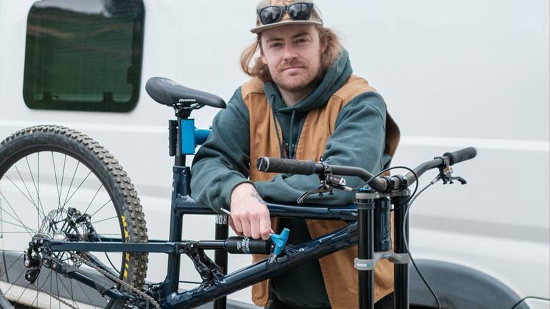 Person vid mountainbike framför en vit skåpbil, iförd keps och solglasögon. Enkla utomhuskläder med en brun väst över en grön hoodie