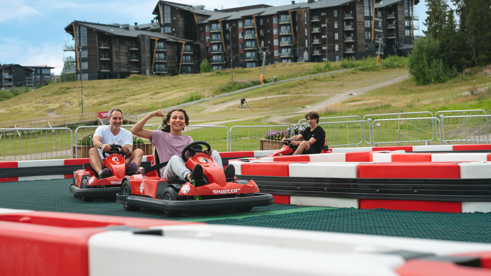 Personer åker gokart på utomhusbana med moderna byggnader i bakgrunden, sommarlandskap
