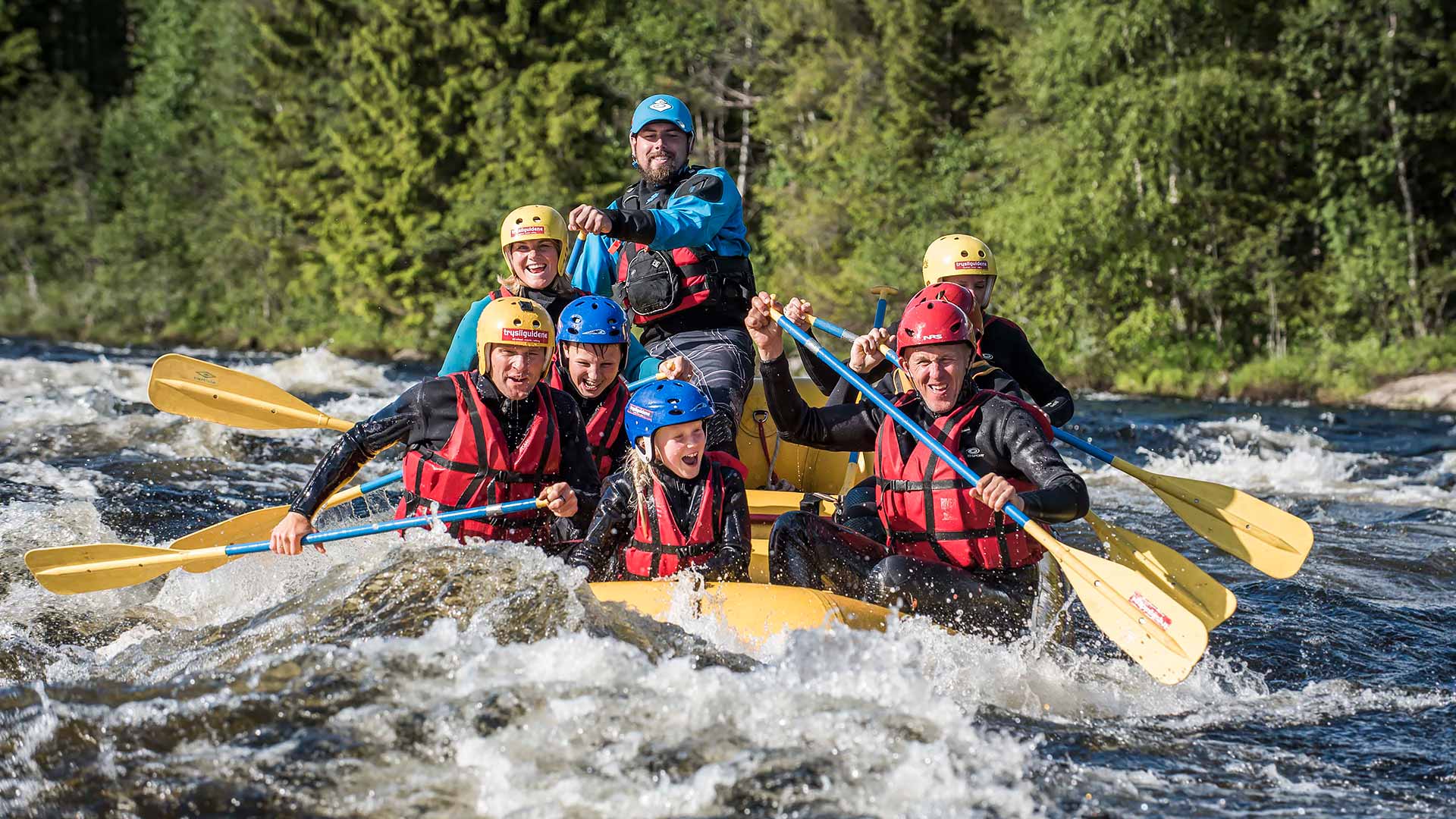 Grupp människor som paddlar en uppblåsbar flotte genom forsande vatten omgivet av skog. De bär flytvästar och hjälmar, och ser koncentrerade ut. Perfekt aktivitet för äventyr och friluftsliv