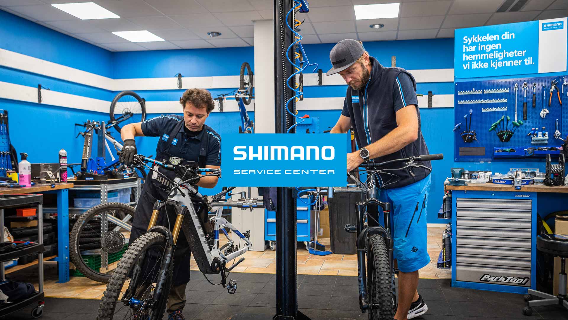 Två cykelmekaniker som arbetar på Shimano Service Center, omgivna av cyklar och verktyg i en professionell verkstad