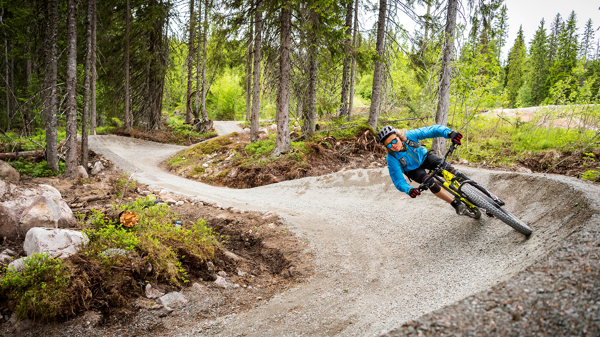 Cyklist i blå jacka cyklar nedför en kurvig grusad led i skogen, utrustad med hjälm, i en grön omgivning