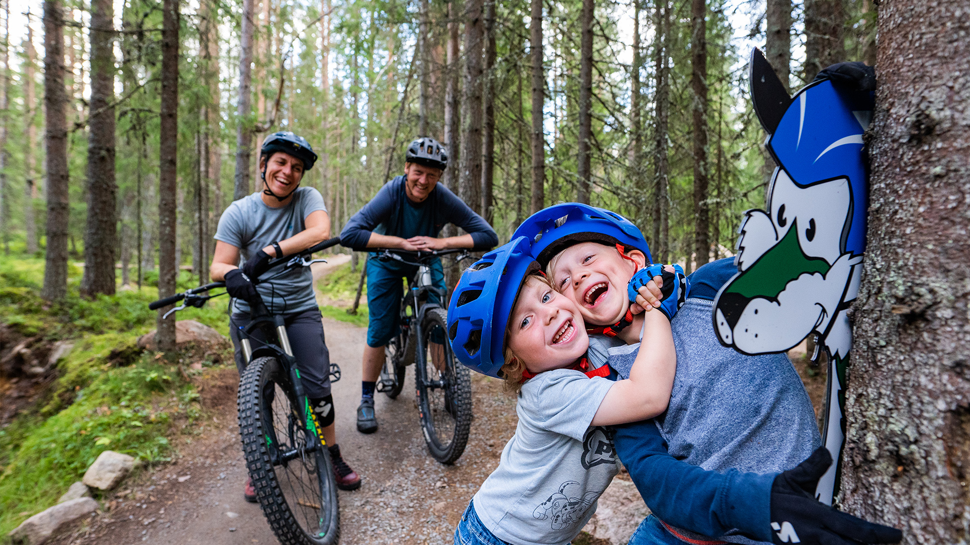 Familj cyklar i skogen på mountainbikeled, två barn med cykelhjälmar leker vid en skylt med en tecknad varg