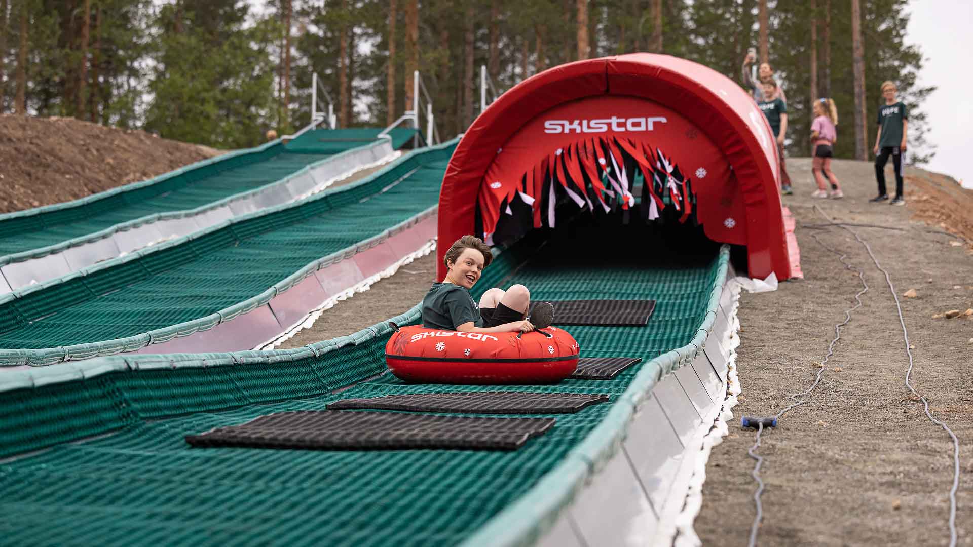 Person glider nerför en rutschkana i en röd tubingring på Skistar-anläggning, omgiven av skog, med leende och skrattande publiken i bakgrunden