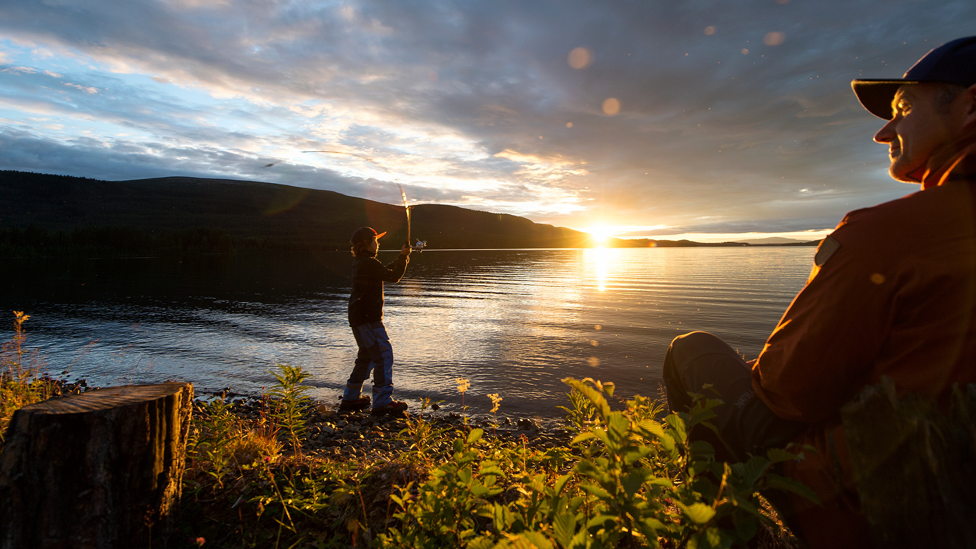 Två personer vid en sjö i solnedgången, en fiskar medan den andra sitter på stranden och njuter av utsikten