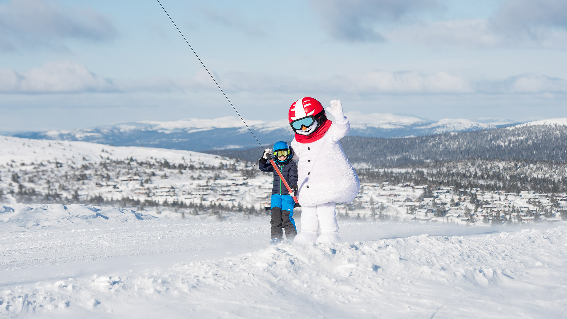 Barn åker skidlift med snögubben Valle i fjällandskap, klara himmel