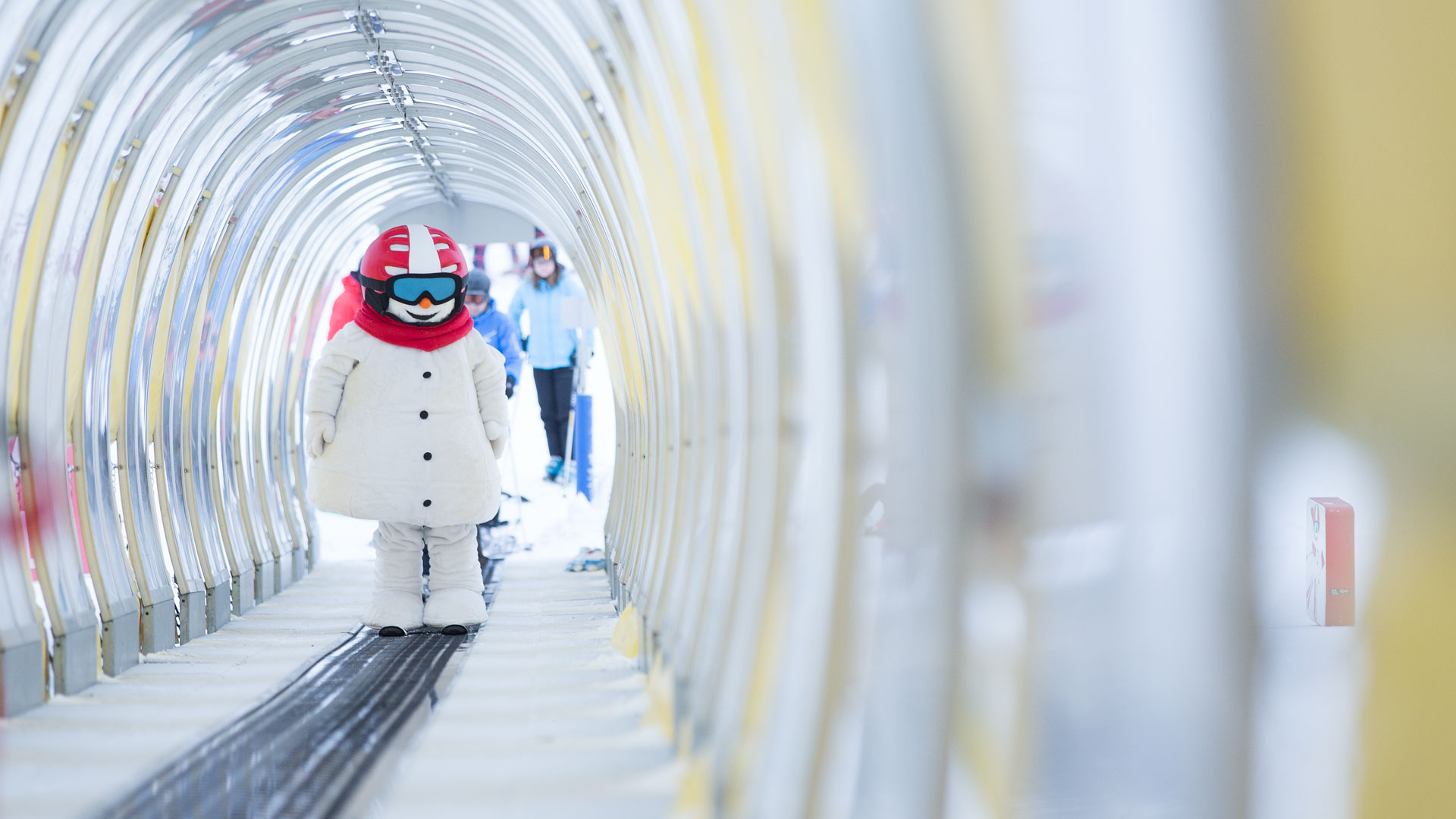 Snögubben Valle med skidglasögon åker på rullband i en täckt gång, med skidentusiaster i bakgrunden. Vinteraktivitet i skidort