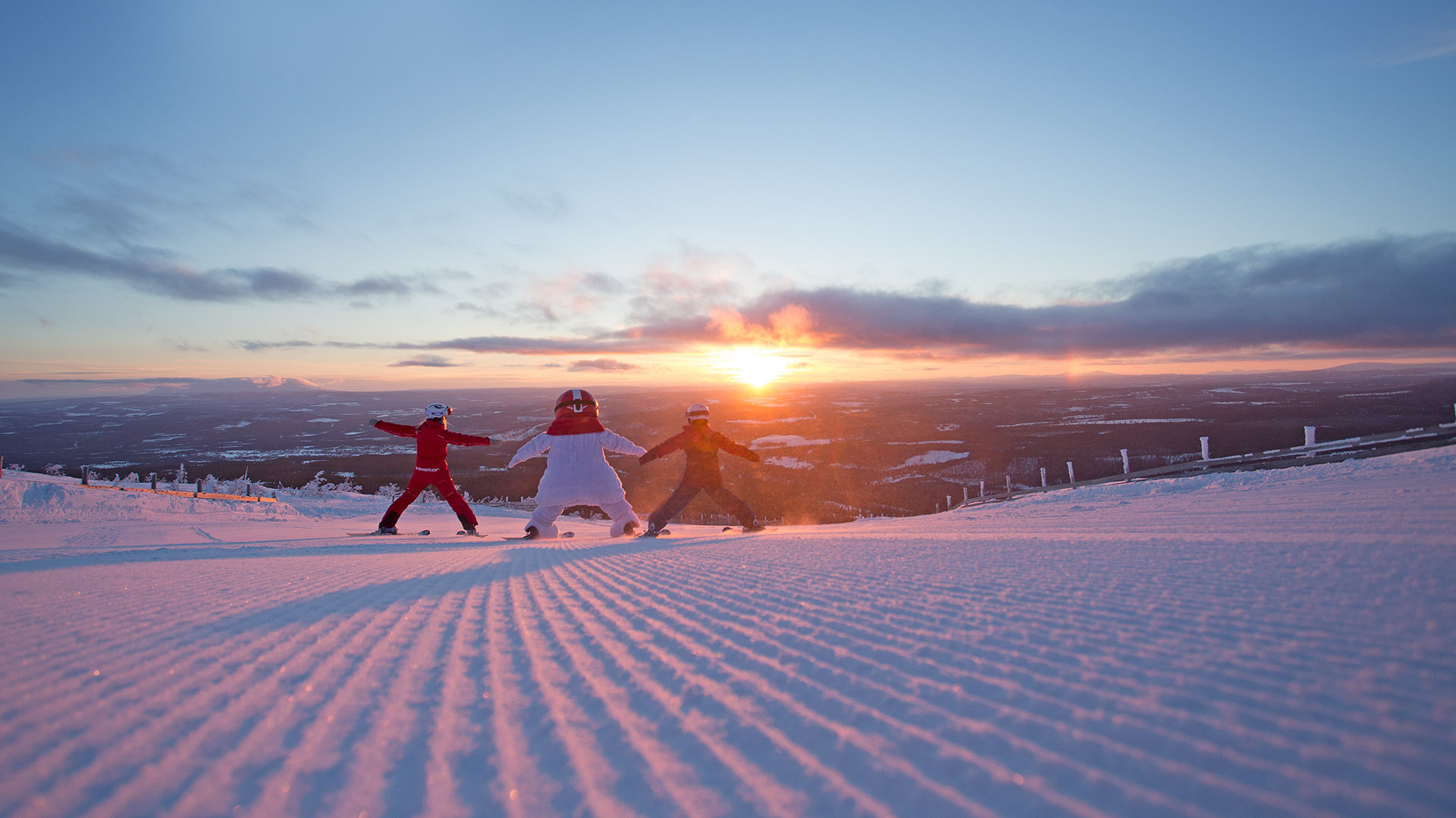 Snögubben Valle och to personer klädda i vinterkläder åker skidor nerför en snötäckt backe vid solnedgången, med ett vidsträckt landskap i bakgrunden