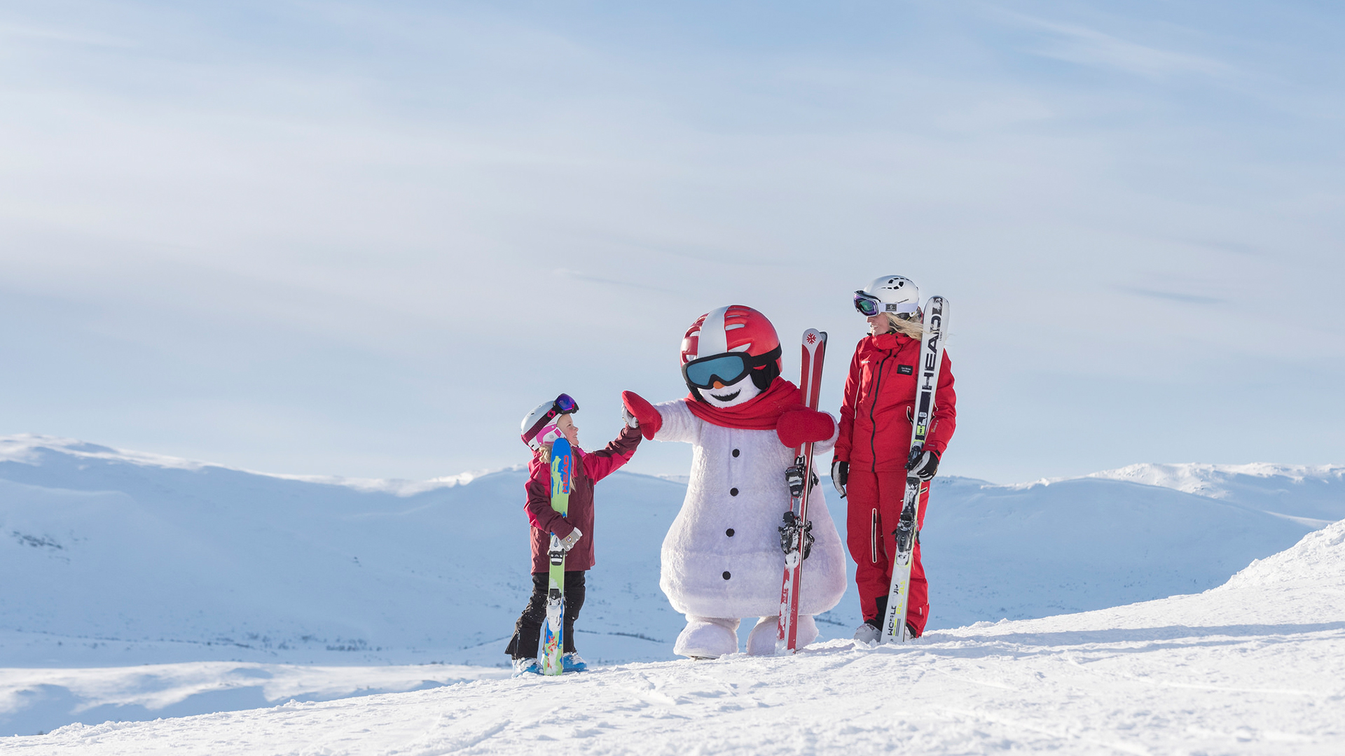 Barn och vuxen i skidkläder bredvid en maskot i form av en snögubbe på en snöig bergstopp