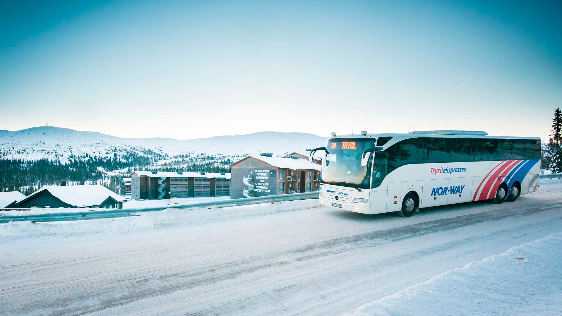 Buss från NOR-WAY Trysilekspressen på en snöig väg med ett vinterlandskap och fjäll i bakgrunden